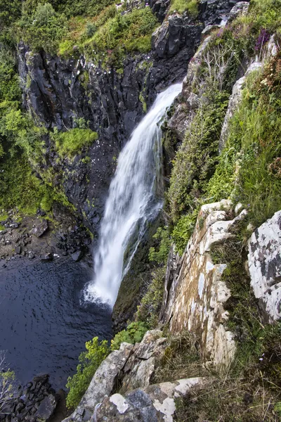 A long exposure photograph of a waterfall flowing down a dark, rocky cliff face covered in green vegetation into a dark pool of water.