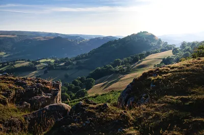A panoramic view of green rolling hills and valleys with a castle ruin on a distant hilltop, bathed in the warm light of the low afternoon sun, with rocky foreground.