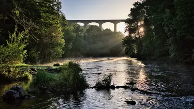 A stone riverbank and flowing water beneath a tall arched aqueduct, framed by trees as sunlight enters from the right.