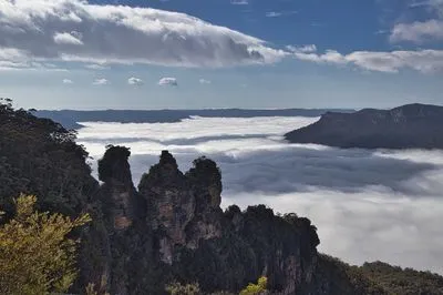 View of The Three Sisters rock formation in the Blue Mountains, Australia, with a dense layer of white clouds filling the valley below and surrounding distant mountain peaks under a blue sky.