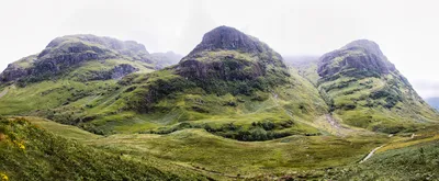 Panoramic view of three large, green, rocky mountains in a valley under a cloudy, misty sky with winding paths.