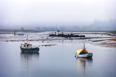 Two boats float on calm water in a misty estuary, with exposed mudflats and the skeletal remains of several boat wrecks in the background.