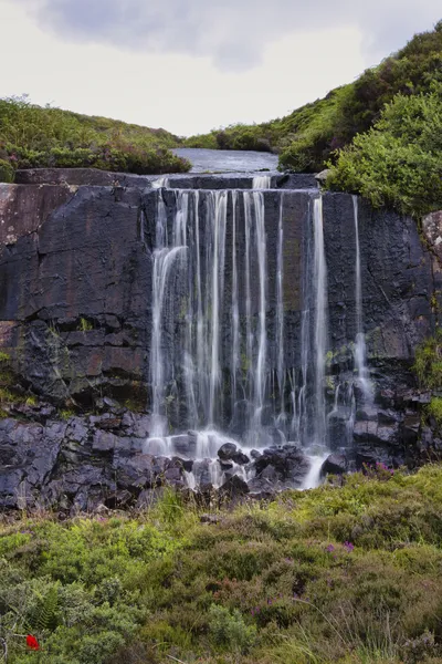 A long exposure photograph of the Kalnakill waterfall cascading over dark rock formations, surrounded by green foliage and purple heather under a cloudy sky.