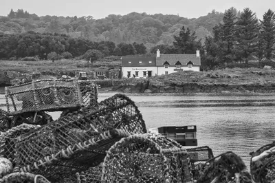 Black and white photograph of a coastal scene with fishing cages in the foreground and a white building named 'The Boathouse' across the water, backed by trees and hills.