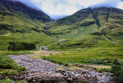 A wide shot of a green mountainous valley with waterfalls flowing down the slopes, a rocky riverbed in the foreground, and a cluster of pine trees.
