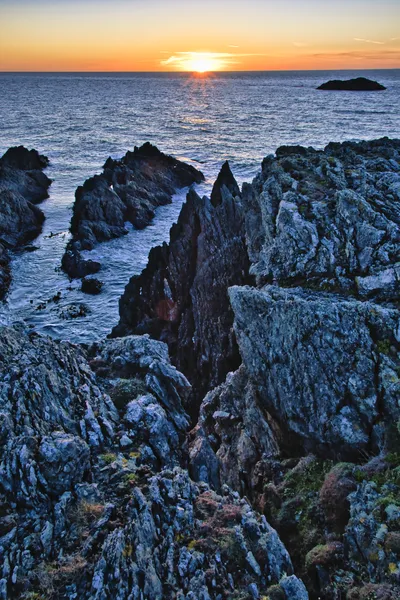 A sunset over a rocky coastline with waves crashing against dark, jagged rocks. The sky is orange and yellow, and the ocean is dark blue. A small island is visible in the distance.