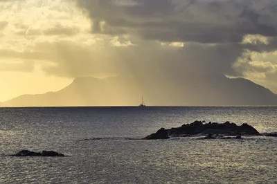 A catamaran sails on the ocean with sun rays breaking through dramatic clouds over distant mountains and islands, with rocks in the foreground.