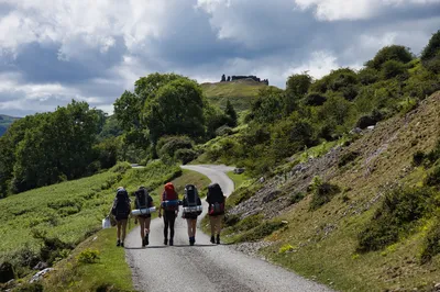 Four hikers with large backpacks walk on a paved road towards a distant hill topped with castle ruins under a cloudy sky.