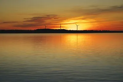 A vibrant sunset over a calm body of water, reflecting orange and red hues. Three wind turbines are silhouetted against the colorful sky on a distant landmass.