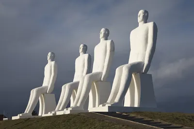 Four massive white statues of seated men, known as Men by the Sea, are positioned on a green grassy mound against a dark, cloudy sky. The figures are looking out towards the horizon.
