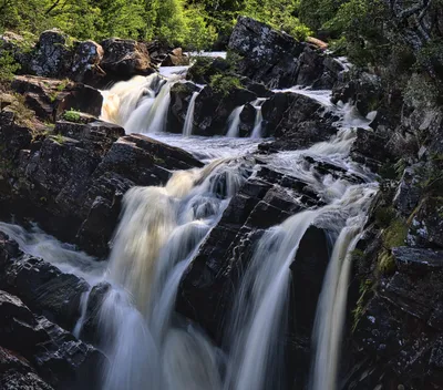 A series of waterfalls with milky white water cascading over dark, layered rocks, surrounded by green trees and foliage.