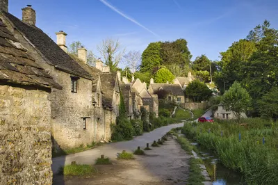 A row of historic stone cottages with slate roofs along a winding road next to a stream, with lush green plants and trees under a blue sky.