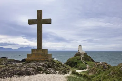 A large stone cross on a rocky hill in the foreground, with a white lighthouse on a small island in the mid-ground. The sea is visible between them, and mountains are in the background under a cloudy 