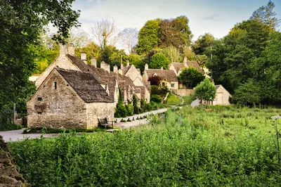 A row of historic stone cottages with slate roofs in a green, tree-lined village, with a grassy field in the foreground.