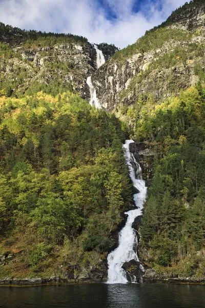 A tall, multi-tiered waterfall flows down a steep, dark rock face covered in green and yellow autumn trees, emptying into a dark body of water at the bottom under a partly cloudy sky.