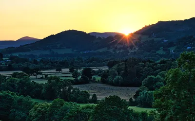 A landscape photograph of a sunset over rolling green hills and valleys, with sunbeams breaking over a distant hilltop.