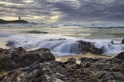 Long exposure photograph of ocean waves flowing over dark rocks on a sandy beach under a cloudy sky, with a distant headland and single tree.