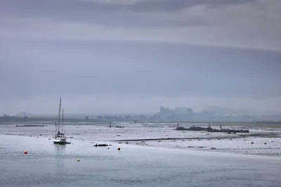 A sailboat and several wooden boat wrecks are visible in the mudflats of a misty estuary under an overcast sky.
