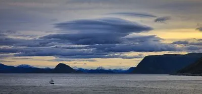 Panoramic view of a fjord with dark, layered clouds overhead and a hint of golden light on the horizon. A small boat moves across the water, with snow-capped mountains and dark hills in the background