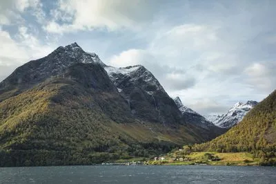 A wide shot of a Norwegian fjord with dark blue water, flanked by steep mountains covered in autumn trees and topped with snow. A small village with red-roofed houses sits on the shore.