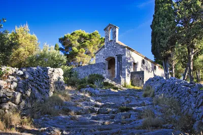 A stone church with a bell archway on a hill, surrounded by green trees and stone walls, with a rocky path leading towards it under a bright blue sky.