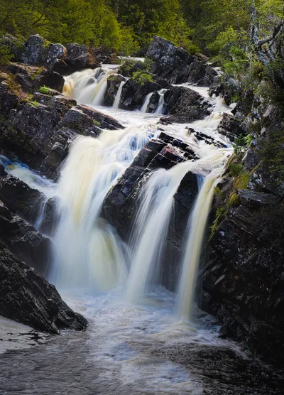 A long exposure photograph of a multi-tiered waterfall flowing over dark, jagged rocks, with lush green trees visible at the top and sides of the gorge.