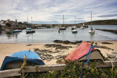 A view of a small harbour with numerous boats moored in calm water, a sandy beach in the foreground, and coastal buildings and hills in the background under a cloudy sky.