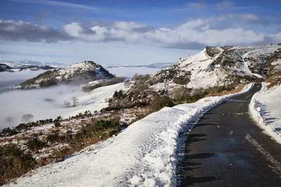 A snowy mountain landscape with a winding road in the foreground. In the distance, Dinas Bran Castle sits on a peak above a valley filled with a thick cloud inversion under a blue sky.