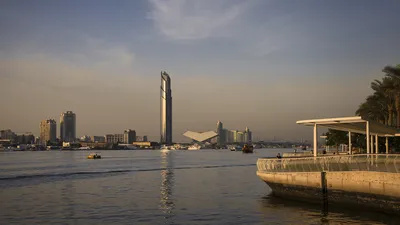 A wide shot of Dubai Creek Harbour at sunset, featuring a tall skyscraper, a modern angular building, boats on the water, and a waterfront promenade with palm trees.