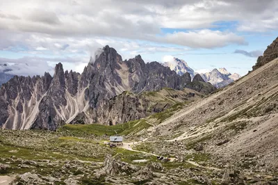 A small mountain hut, Rifugio Lavaredo, is nestled in a green valley surrounded by dramatic, jagged peaks of the Dolomites under a cloudy sky with patches of blue.
