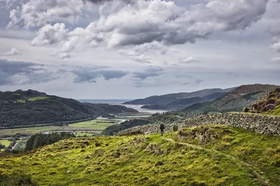 A person walks on a narrow path on a grassy hillside, next to a stone wall. In the background, a wide river estuary winds through a valley with forested hills and distant mountains under a cloudy sky.