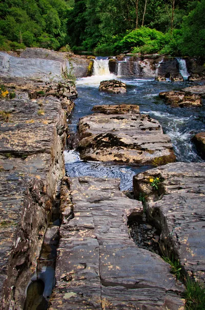 A river with multiple waterfalls flowing over dark, layered rocks, surrounded by dense green forest under a bright sky.