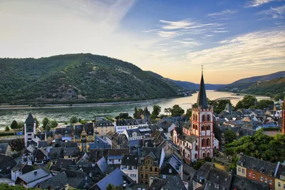 An elevated view of the historic town of Bacharach, Germany, at sunset. A prominent red and white church steeple rises above traditional buildings, with the Rhine River and forested hills in the backg