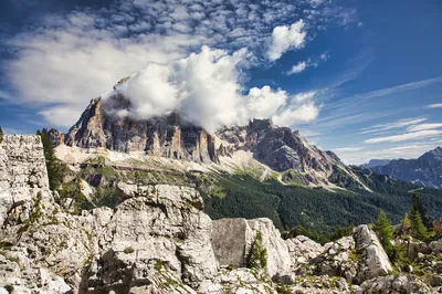 A panoramic view of rugged, light-colored mountains with green forests and white clouds partially obscuring the peaks under a blue sky.