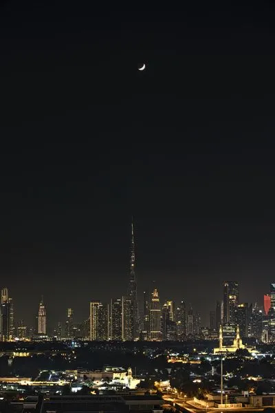 A vertical night shot of the Dubai skyline, dominated by the brightly lit Burj Khalifa skyscraper. A crescent moon is visible in the dark sky above the city lights.