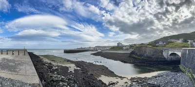 Panoramic view of a coastal town with a long concrete pier extending into the sea, a river estuary, a rocky beach, and houses on green hills under a cloudy sky.