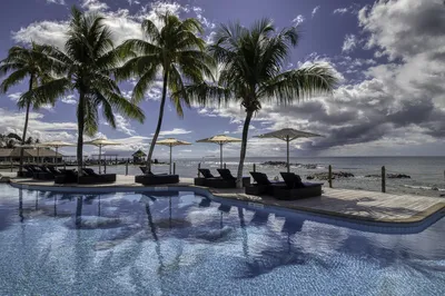 A resort swimming pool with blue water, surrounded by palm trees, sun loungers, and umbrellas, with a rocky beach and ocean under a cloudy sky in the background.