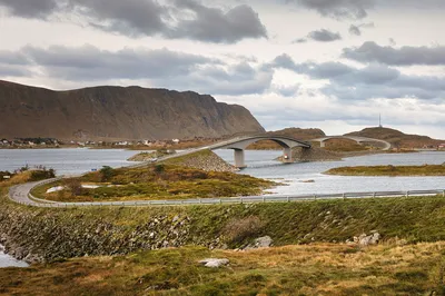 A landscape view of a winding road and two arched bridges spanning across water, connecting small islands. Mountains rise in the background under a cloudy sky, with a village visible along the distant