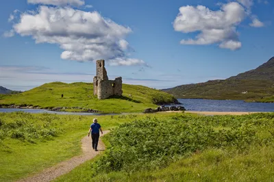 A person walks on a dirt path towards the ruins of Ardvreck Castle, a stone structure on a green hill next to a blue lake, with mountains and a cloudy sky in the background.