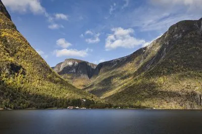 A wide shot of a Norwegian fjord with a small village at the base of steep, tree-covered mountains displaying autumn colors under a blue sky with white clouds.