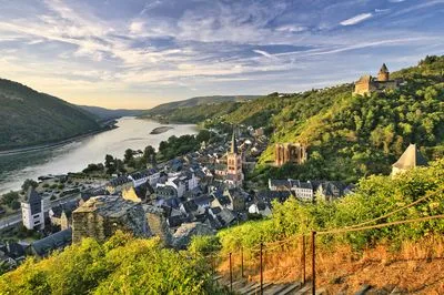 Panoramic view of Bacharach, Germany, with the Rhine River flowing through a valley. The town features a prominent church, hillside ruins, and a castle, all bathed in warm sunset light.
