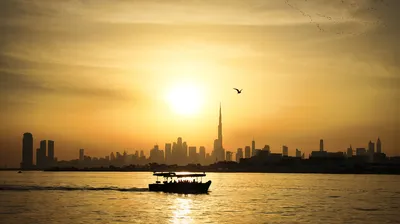 A silhouette of a dhow boat with passengers on water, with the Dubai skyline and Burj Khalifa in the background against a golden sunset sky. A bird flies above the skyscraper.