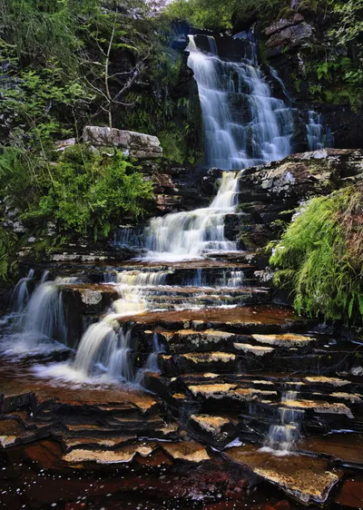A multi-tiered waterfall cascades over dark, layered rocks surrounded by lush green vegetation, captured with a long exposure effect.