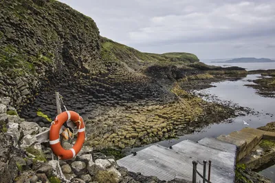 A coastal scene featuring dark hexagonal basalt columns forming cliffs and a rocky shore, with an orange lifebuoy on a wooden post and a small jetty.