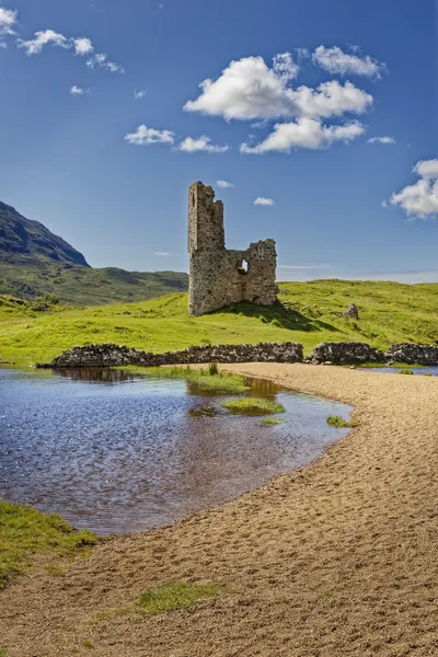 The ruins of Ardvreck Castle stand on a grassy hill next to a body of water with a sandy bank under a blue sky with white clouds.