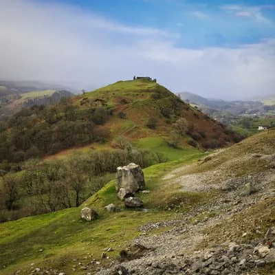 Dinas Bran Castle ruins on a green hill with a winding path, surrounded by rolling hills and trees under a partly cloudy sky.