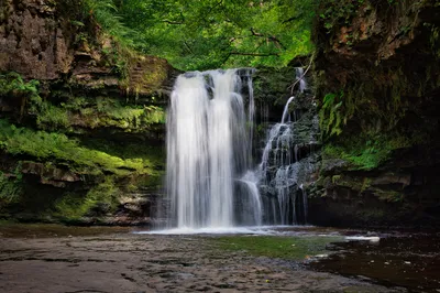 A long exposure photograph of a waterfall with white, silky water flowing over dark, moss-covered rocks into a shallow pool, surrounded by green foliage and trees.