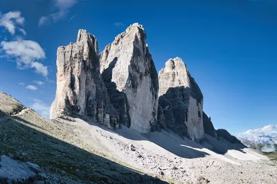 A panoramic view of three large, rugged mountain peaks with a winding hiking trail and small figures of hikers on the left, under a bright blue sky with scattered clouds.