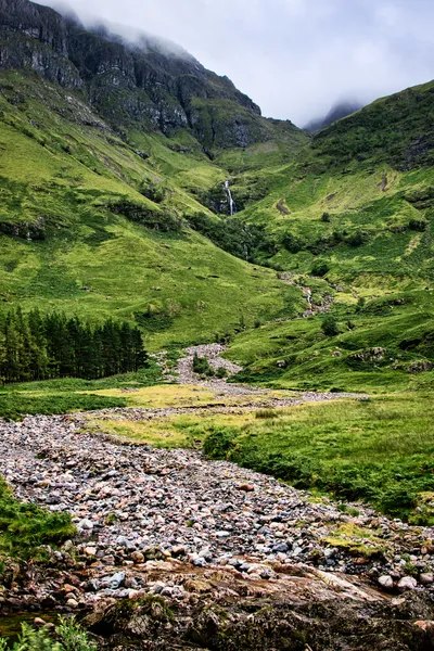 A portrait photograph of a lush green valley in Scotland, featuring steep mountains, a prominent waterfall, and a rocky stream bed with scattered trees.
