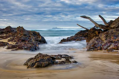 A long exposure photograph of waves washing over a rocky beach with sand and large pieces of driftwood under a cloudy sky. A distant coastline is visible.